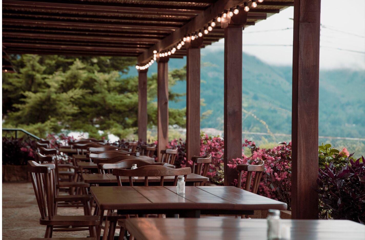 An outdoor dining area with wooden tables and chairs, string lights, and a view of mountains.