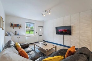 A modern living room with a sofa, a coffee table, a television displaying 'NETFLIX,' and a bookshelf. - 2BR House Sleeps 5 | Near Centre Gloucester (Gloucestershire)