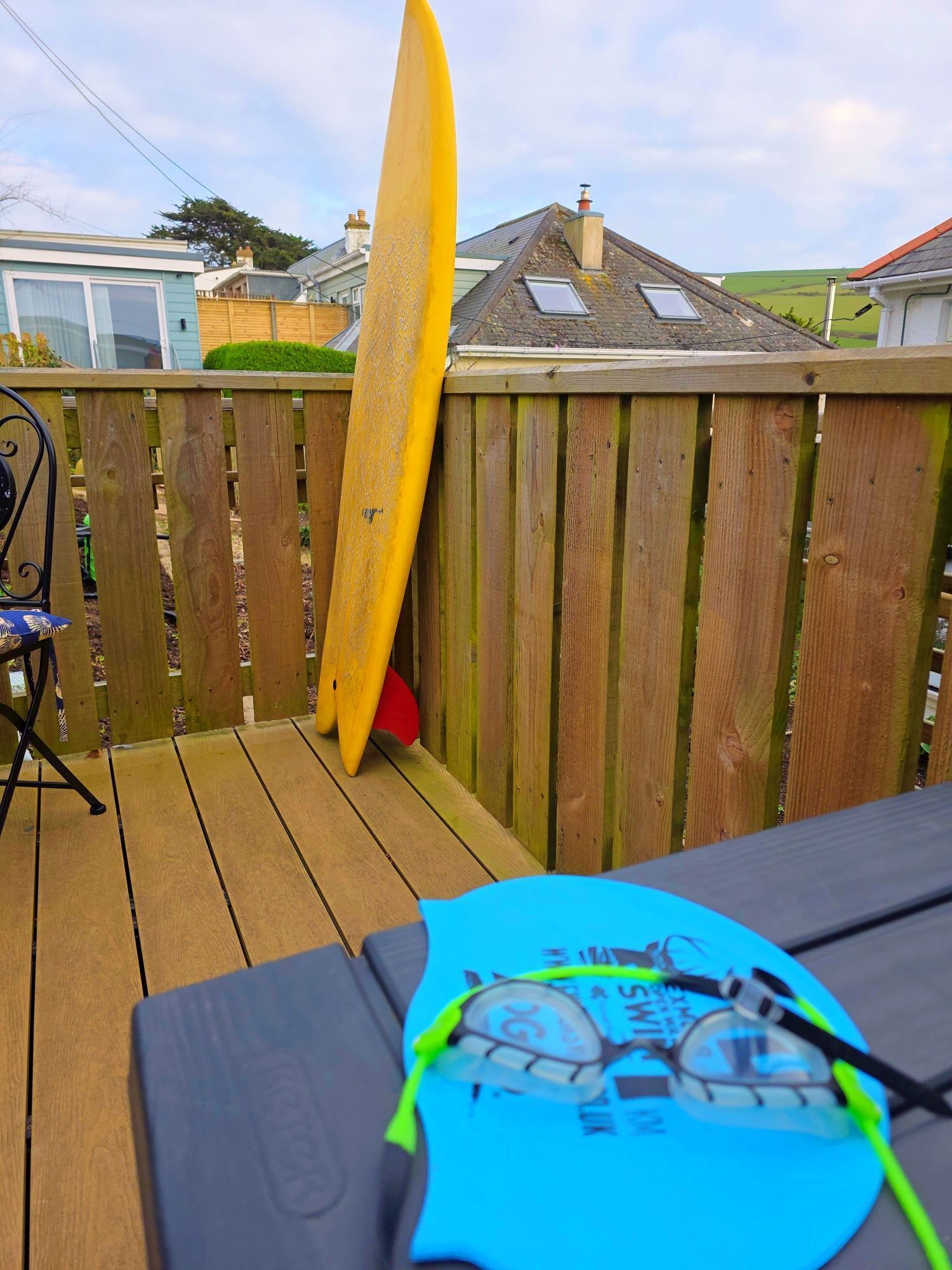 A wooden deck with a surfboard leaning against a wooden fence, a table with a blue shirt and sunglasses, and a house in the background.