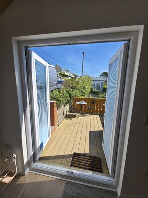 Blick durch ein Fenster auf einen Raum mit einer Holzterrasse, einem Tisch mit Stühlen und einer Ziegelwand.