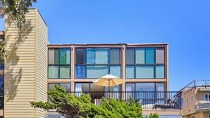 Un edificio moderno con ampie finestre, un balcone con sedia e ombrellone, e un albero di fronte.