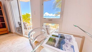 Ett rum med ett glasbord där konstverk visas, en stol och utsikt över en strand genom en skjutdörr.