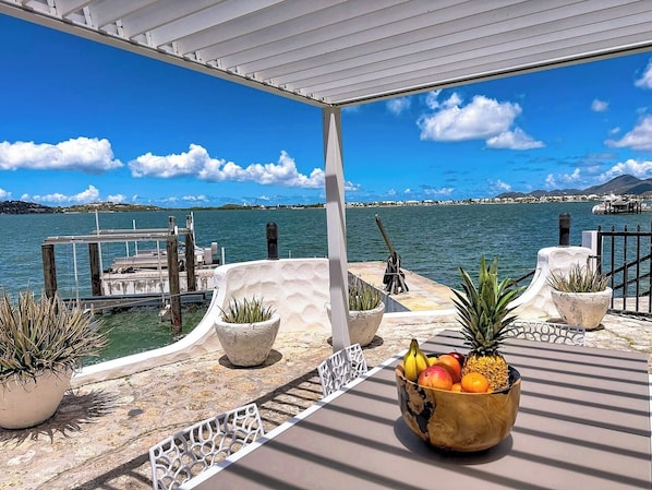 A covered outdoor area with potted plants and a fruit bowl, overlooking a body of water and dock.