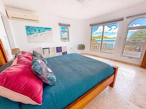 A bedroom with a bed, a desk, a chair, and large windows with a view.