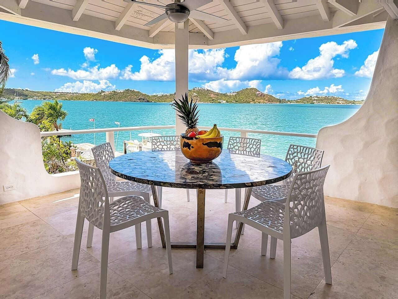 A covered outdoor dining area with a round table and white chairs, overlooking a body of water and hills.