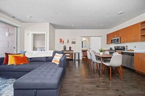 Un salon moderne comprenant un canapé d’angle, un coin repas, une cuisine et une porte visible donnant sur une autre pièce.