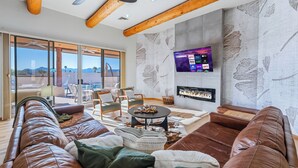 Ein modernes Wohnzimmer mit Ledersofas, einem Kamin, einem Fernseher und großen Fenstern mit Blick auf eine gebirgige Landschaft.