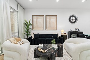 A modern living room with a black sofa, white armchairs, a glass coffee table, and framed artwork on the wall. - New Luxury Dallas Skyline+Rooftop+Fireplace+Garage+BishopArts (Dallas)