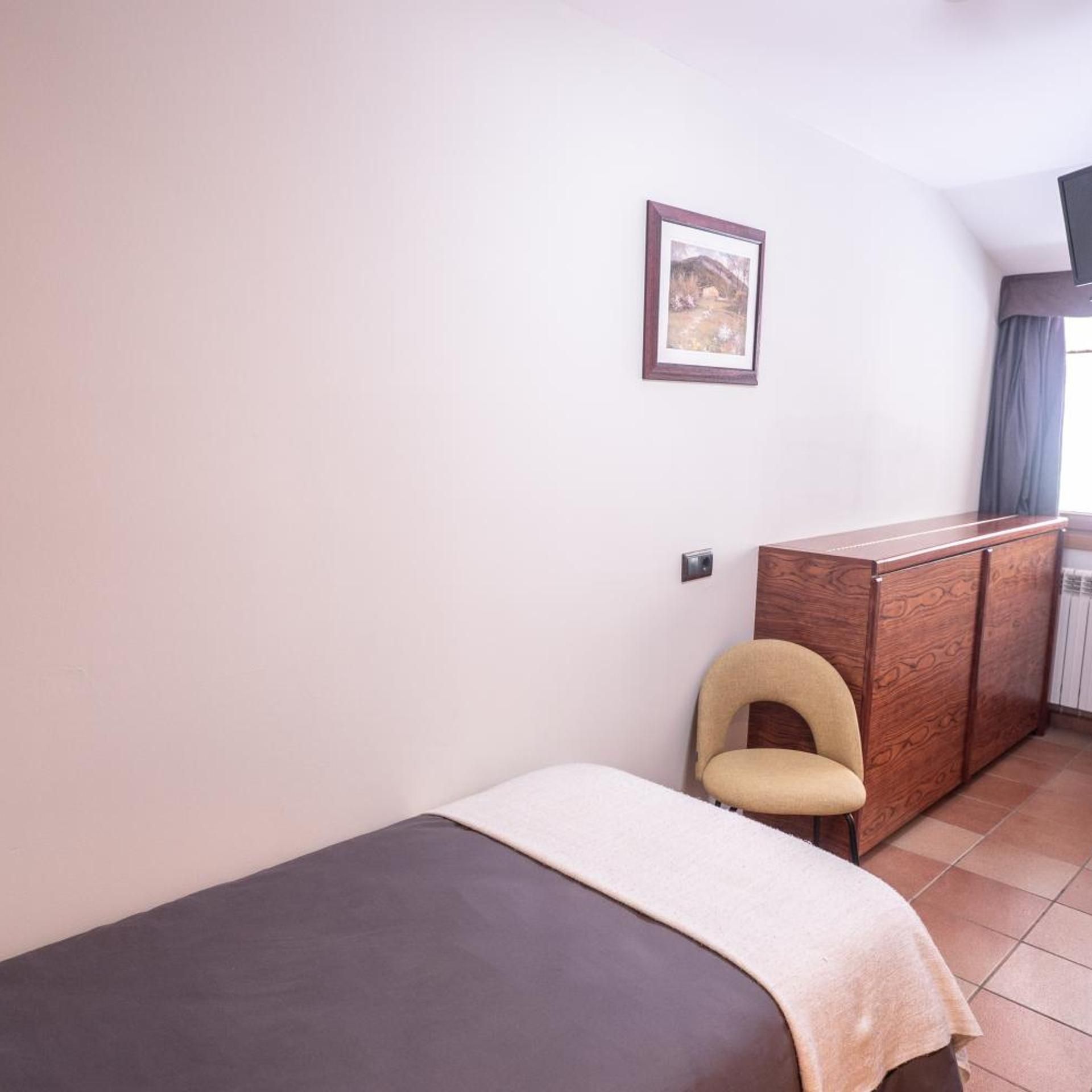 A small, simple room with a single bed, a wooden cabinet, a chair, and a wall-mounted picture.