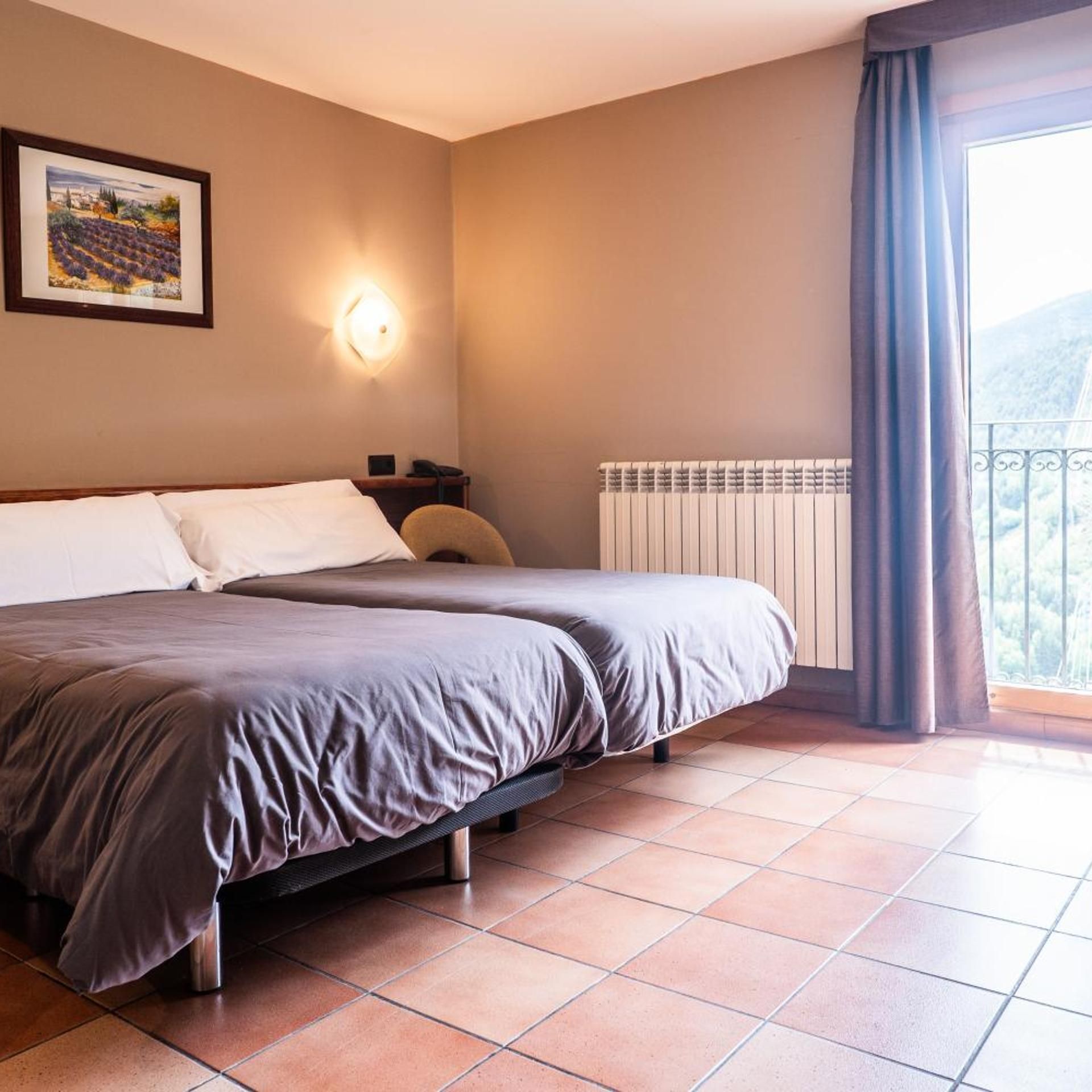 A hotel room with a bed, a nightstand, a wall-mounted light, a radiator, and a window with curtains.