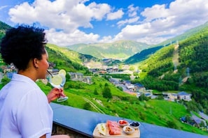 Una persona sosteniendo un vaso con bebida, contemplando un valle con edificios y vegetación.
