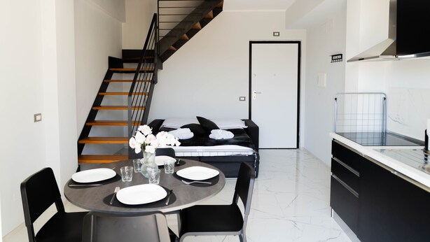 Een moderne keuken met een zwart aanrecht, een eethoek met een ronde tafel, en een trap naar een slaapkamer.