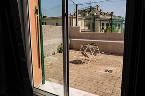 Blick durch ein Fenster auf eine Pflasterterrasse mit einer Metallleiter und einem grünen Zaun.