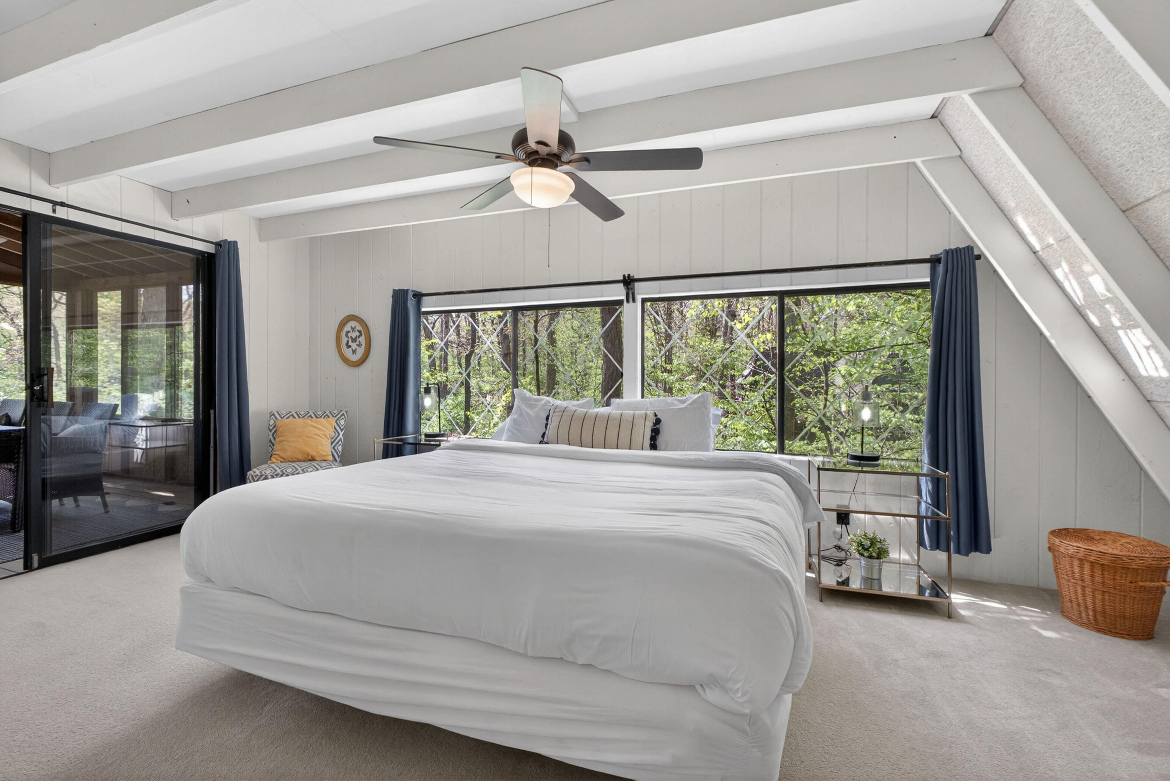 Une chambre dotée d’un grand lit, d’un ventilateur de plafond, d’une porte-fenêtre coulissante donnant sur un espace extérieur et offrant une vue sur les arbres à travers les fenêtres.