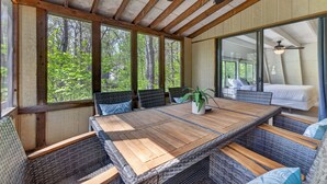 A dining area with a wooden table and wicker chairs, large windows with a forest view, and a ceiling fan.