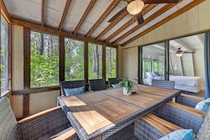 Ein Essbereich mit Holztisch und Korbstühlen, großen Fenstern mit Blick auf einen Wald und einem Deckenventilator.
