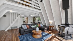 A modern loft with a staircase, a black leather sofa, a round wooden coffee table, and a wood-burning stove.