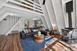A modern loft with a staircase, a black leather sofa, a round wooden coffee table, and a wood-burning stove.