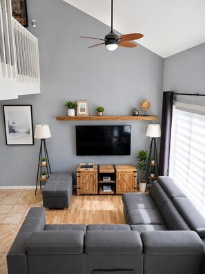 A modern living room with a sectional sofa, a flat-screen TV, wooden shelves, a ceiling fan, and a floor lamp. - Le Chalet De La Capitale (Beaupré)