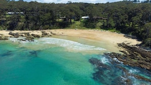 A coastal landscape with clear turquoise waters, sandy beach, and dense forested hills.