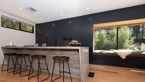 A modern kitchen with a bar area, wooden flooring, and large windows offering a view of trees.