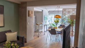 Ein modernes Wohnzimmer mit einer Couch, einem Essbereich mit Tisch und Stühlen, einem Fernseher und einer Küchenzeile mit Wagen und Pflanzen.