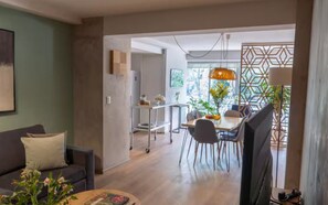 Una sala de estar moderna con un sofá, un comedor con mesa y sillas, un televisor y una zona de cocina con un carrito y plantas.