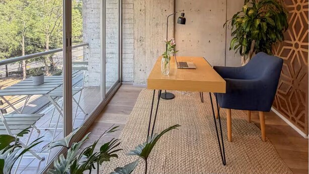 Ein modernes Zimmer mit Holztisch, blauem Stuhl, Topfpflanze und Blick auf Bäume durch große Fenster.
