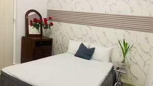 Een slaapkamer met een bed, een kaptafel, een spiegel, een vaas met rode bloemen en een klein tafeltje met een plant.