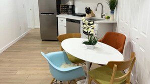 Cocina compacta con armarios blancos, microondas y refrigerador. Hay una mesa de comedor con dos sillas, una azul y otra verde.