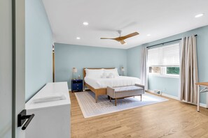 Ein Schlafzimmer mit Bett, Nachttischen, Deckenventilator, Fenster mit Vorhängen und Blick ins Freie.