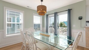 A dining area with a glass table and white wicker chairs, large windows with blinds, and a woven pendant light. - Livin' Easy | 4 Bedroom House with new Private Pool walking distance to Beach and Restaurants (Holden Beach)
