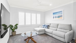 A modern living room with a grey sofa, a glass coffee table, and a ceiling fan. - Casa Azul - Sun, Swim & Coastal Ease (Burrill Lake)