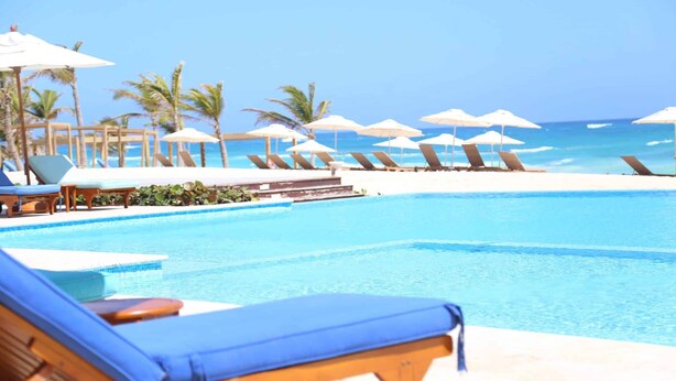 A poolside area with lounge chairs and umbrellas, a clear view of the ocean, and a sandy beach in the background. - Amazing Condo infinity Pool & Golf View at Cana Rock Condos (Punta Cana)