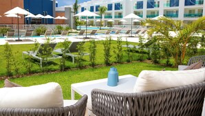 A pool area with lounge chairs and umbrellas, a white table with a blue vase, and a building with multiple balconies. - Amazing Condo infinity Pool & Golf View at Cana Rock Condos (Punta Cana)