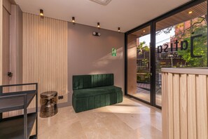 Un espace d’accueil avec un canapé vert, un comptoir de réception en bois, une porte vitrée donnant sur l’extérieur et un panneau indiquant la présence d’un balcon.