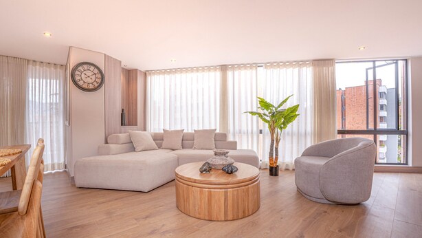 Ein modernes Wohnzimmer mit einem Ecksofa, einem runden Couchtisch, einem grauen Sessel und einem großen Fenster mit Vorhängen.