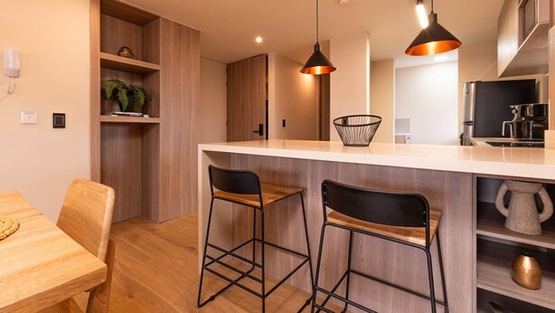 Eine moderne Küche mit Holzschränken, einer weißen Arbeitsplatte, zwei Barhocker, Pendelleuchten und einem Einbauofen.