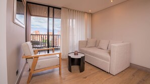 Ein modernes Wohnzimmer mit Sofa, Sessel, kleinem Tisch und großem Fenster mit Blick auf Gebäude und Berge.