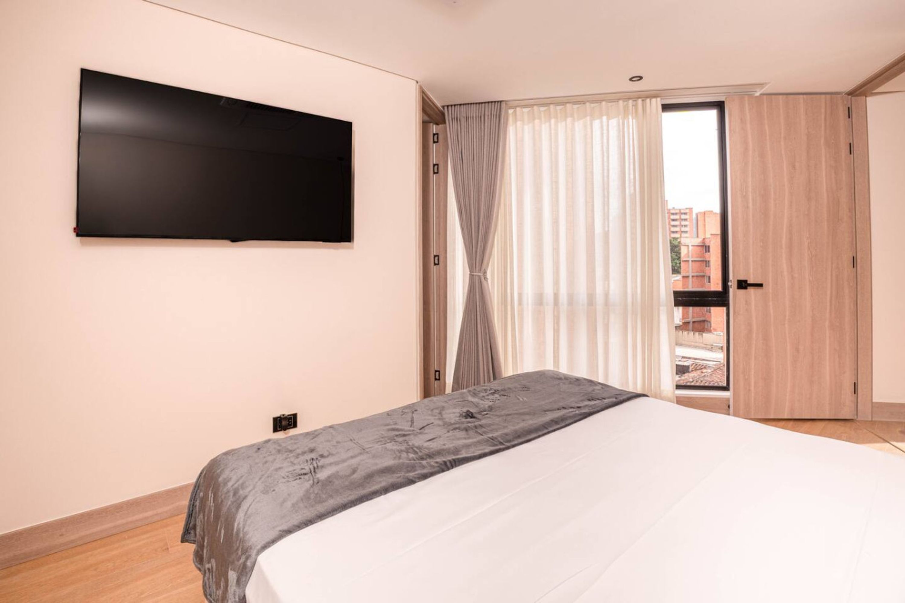 Une chambre à coucher avec un lit, un téléviseur fixé au mur, une porte coulissante et une vue sur les bâtiments à l’extérieur.