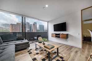 Ein modernes Wohnzimmer mit einem großen Fenster, einer Couch, einem Couchtisch mit einem Buch, einem an der Wand montierten Fernseher und einer kleinen Pflanze.