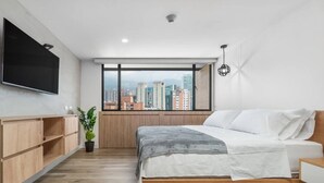 A modern bedroom with a bed, a TV, a window with a city view, and a plant. - Go Living Suites - 1205 (Medellín)