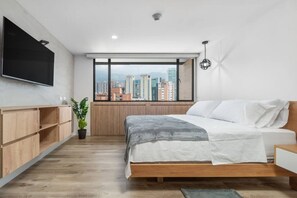 A modern bedroom with a bed, a TV, a window with a city view, and a plant. - Go Living Suites - 1205 (Medellín)