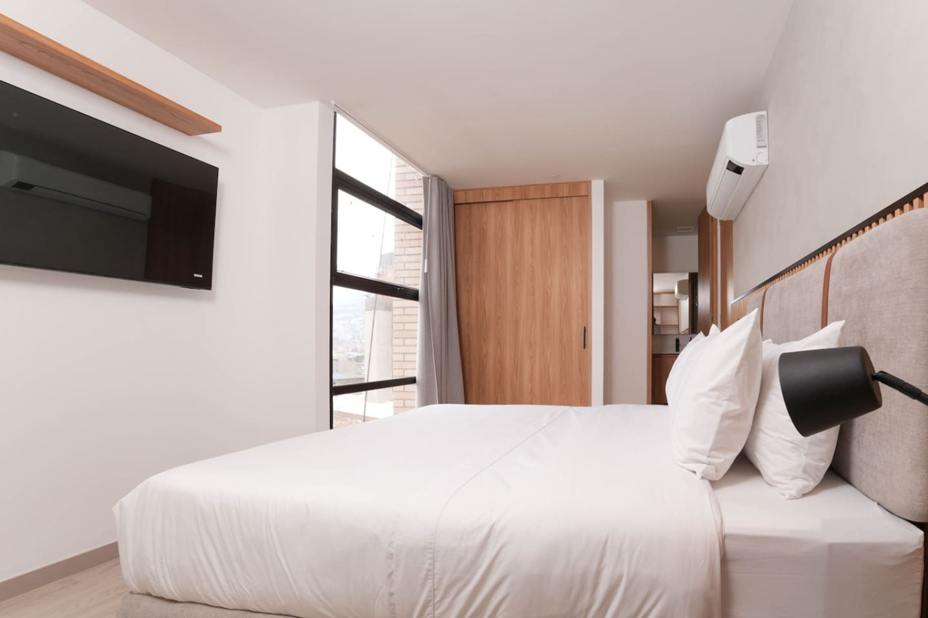 A modern bedroom with a bed, a mounted TV, an air conditioning unit, and a view through a window.