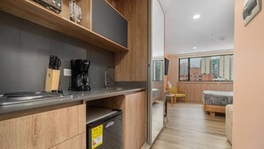 A compact kitchen with a refrigerator, coffee maker, and knife block. A sliding door leads to a bedroom with a bed and a chair. - Go Living Suites - 1207 (Medellín)
