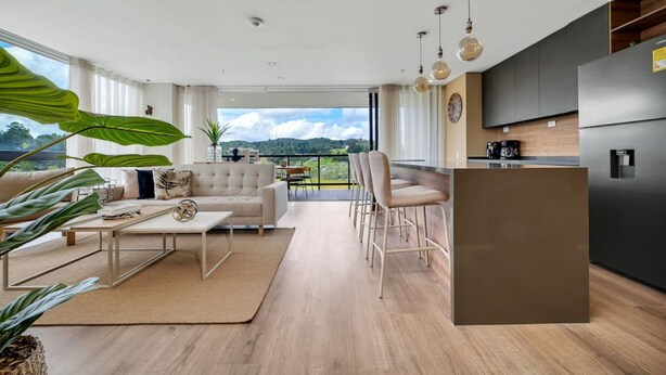A modern living room with a sofa, coffee table, and chairs, a kitchen with a refrigerator and stove, and a large window with a view. - Olive Country Living - 301 (Envigado)