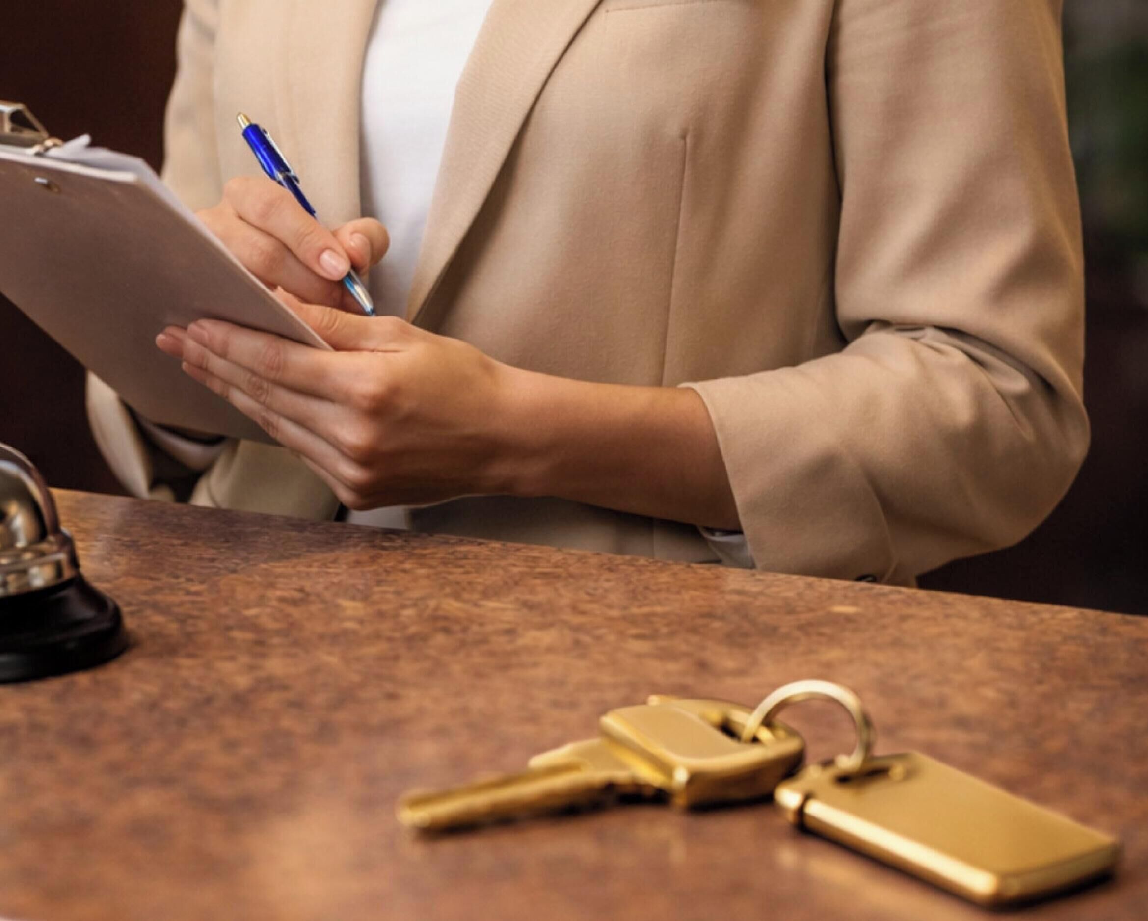 A person in a beige blazer holding a clipboard and pen, with keys and a keychain on a wooden surface.