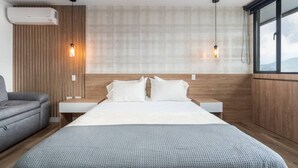Ein modernes Hotelzimmer mit einem hölzernen Kopfteil, einem Bett mit weißen Leinen, einem grauen Sofa und zwei Pendelleuchten.