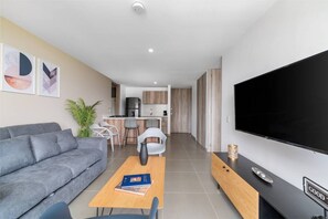 Un salon moderne avec un canapé gris, une table basse en bois, un téléviseur et une cuisine en arrière-plan.