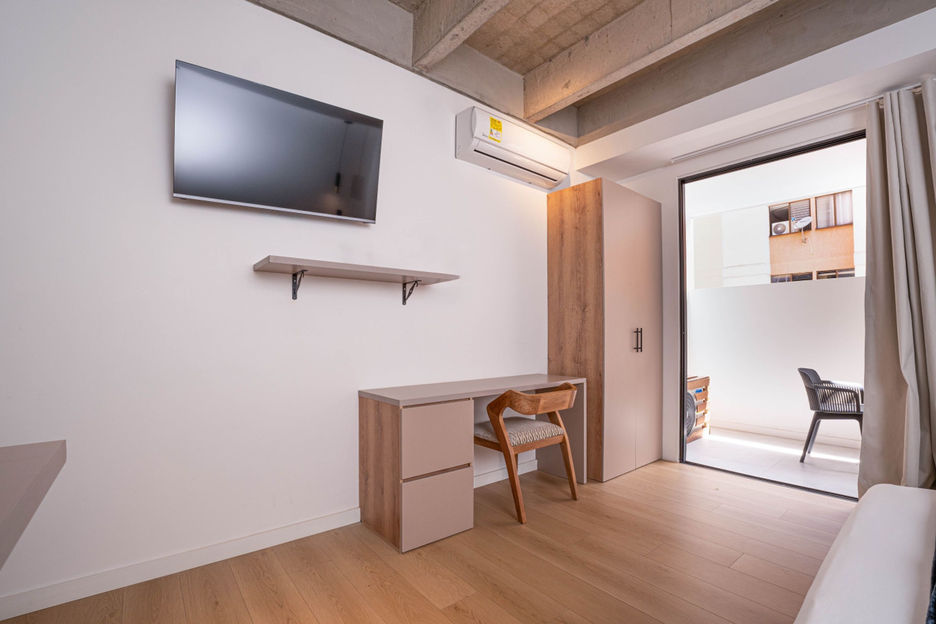 Habitación moderna con televisión de pantalla plana, aire acondicionado integrado, un escritorio de madera, una silla y una puerta corrediza de vidrio que da a un balcón.